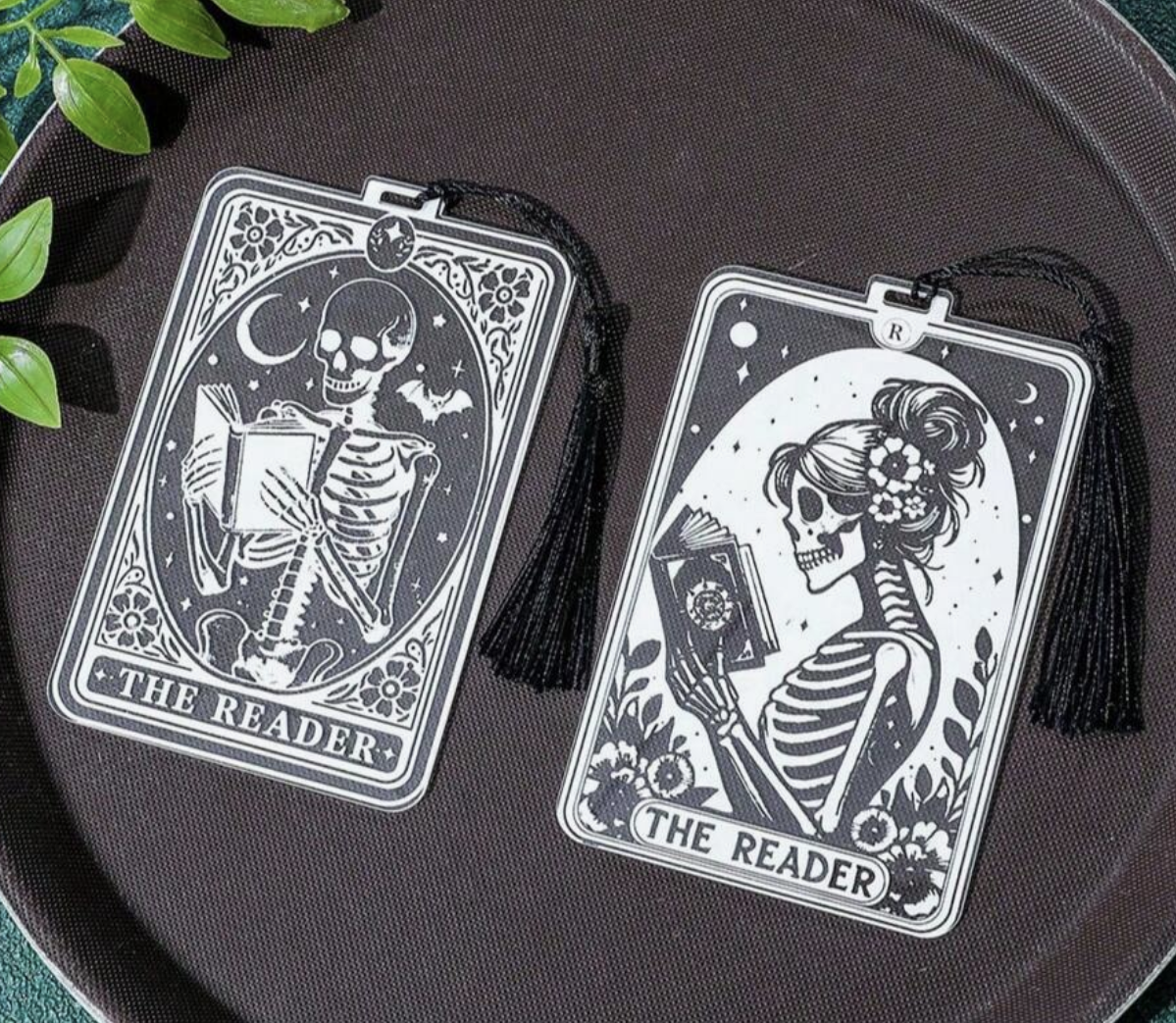 Two skeleton-themed bookmarks labeled 'The Reader' on a dark surface with green leaves.