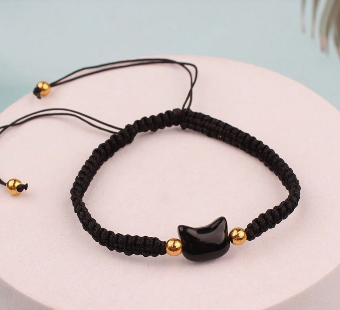 Black macrame bracelet with gold beads and a black cat-shaped charm on a light blue background.