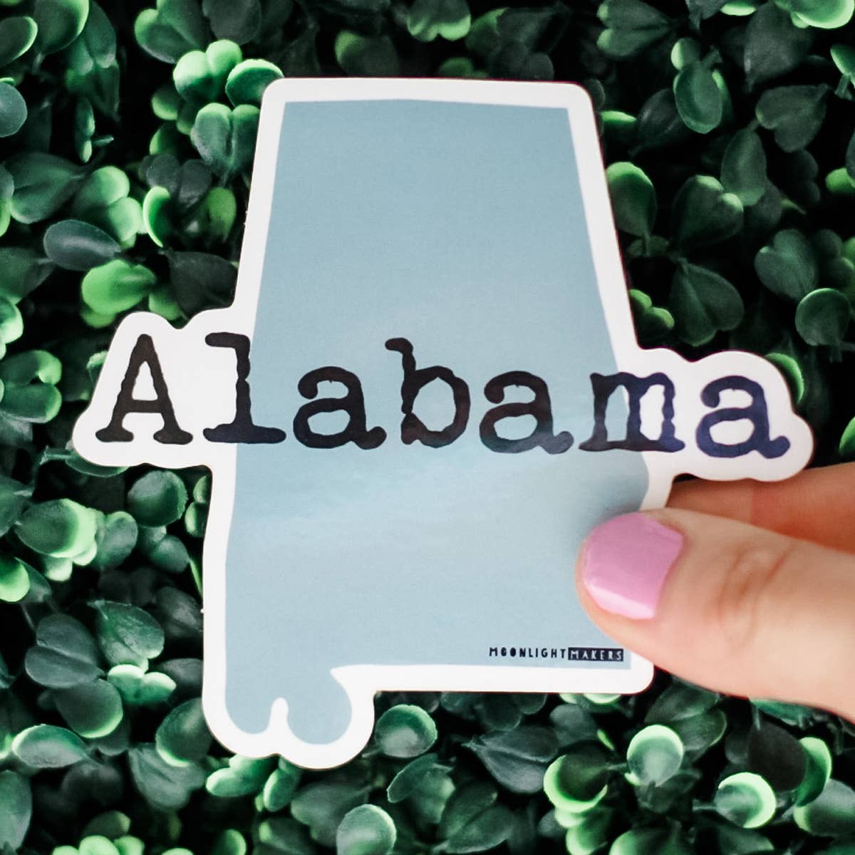 Alabama state outline vinyl sticker with waterproof and weatherproof finish held over green leafy background