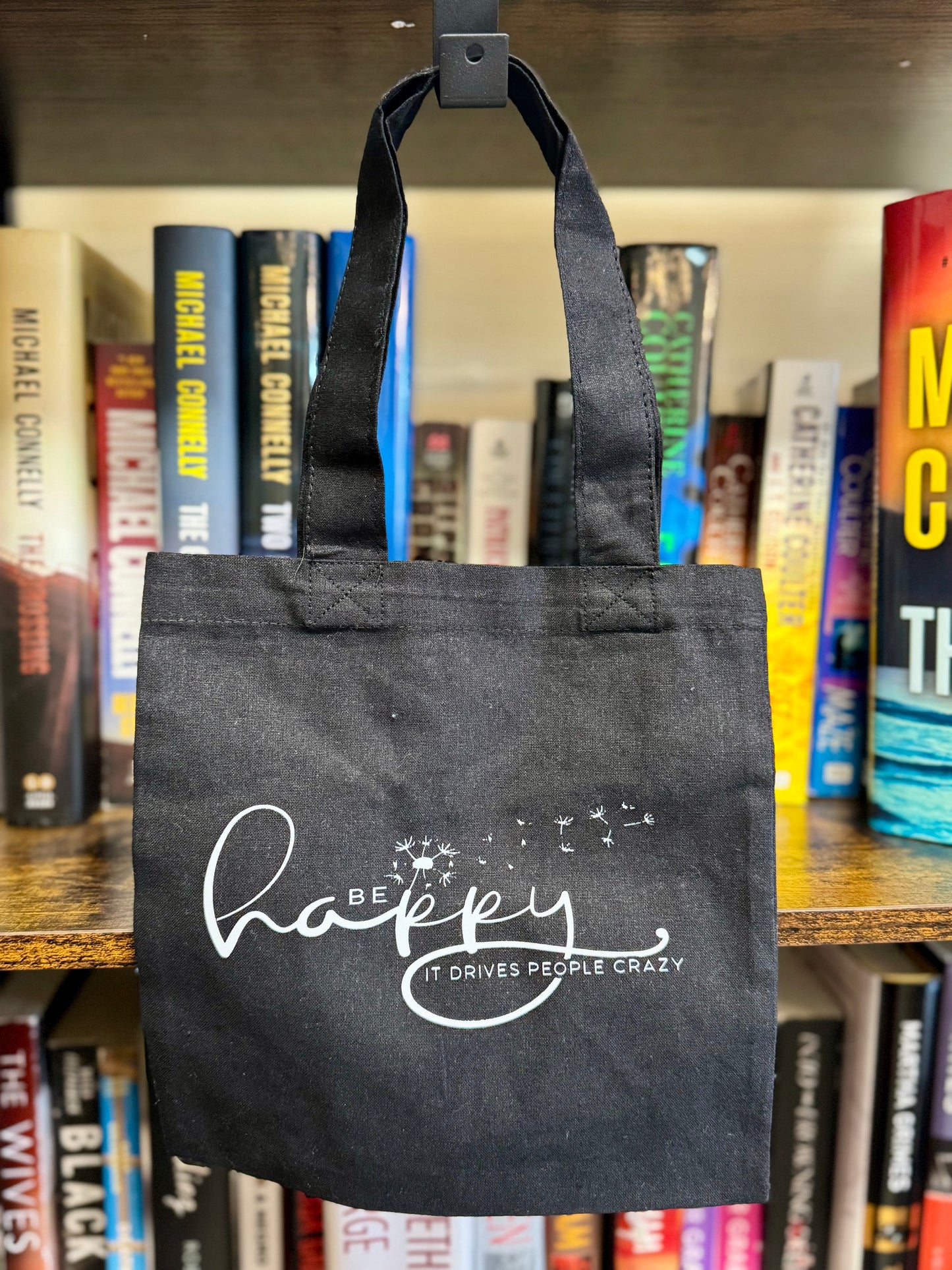 Small natural canvas tote bag with Be Happy It Drives People Crazy and a dandelion being blown to make a wish on one side, and The Burrow Bookstore on the other side.
