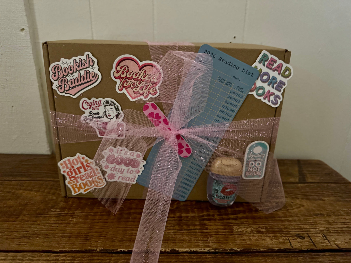 Blind Date with a Book romance-themed gift box wrapped in kraft paper and pink tulle ribbon, decorated with bookish stickers, reading list bookmark, nail file, and small jar accessory, displayed on wooden surface.