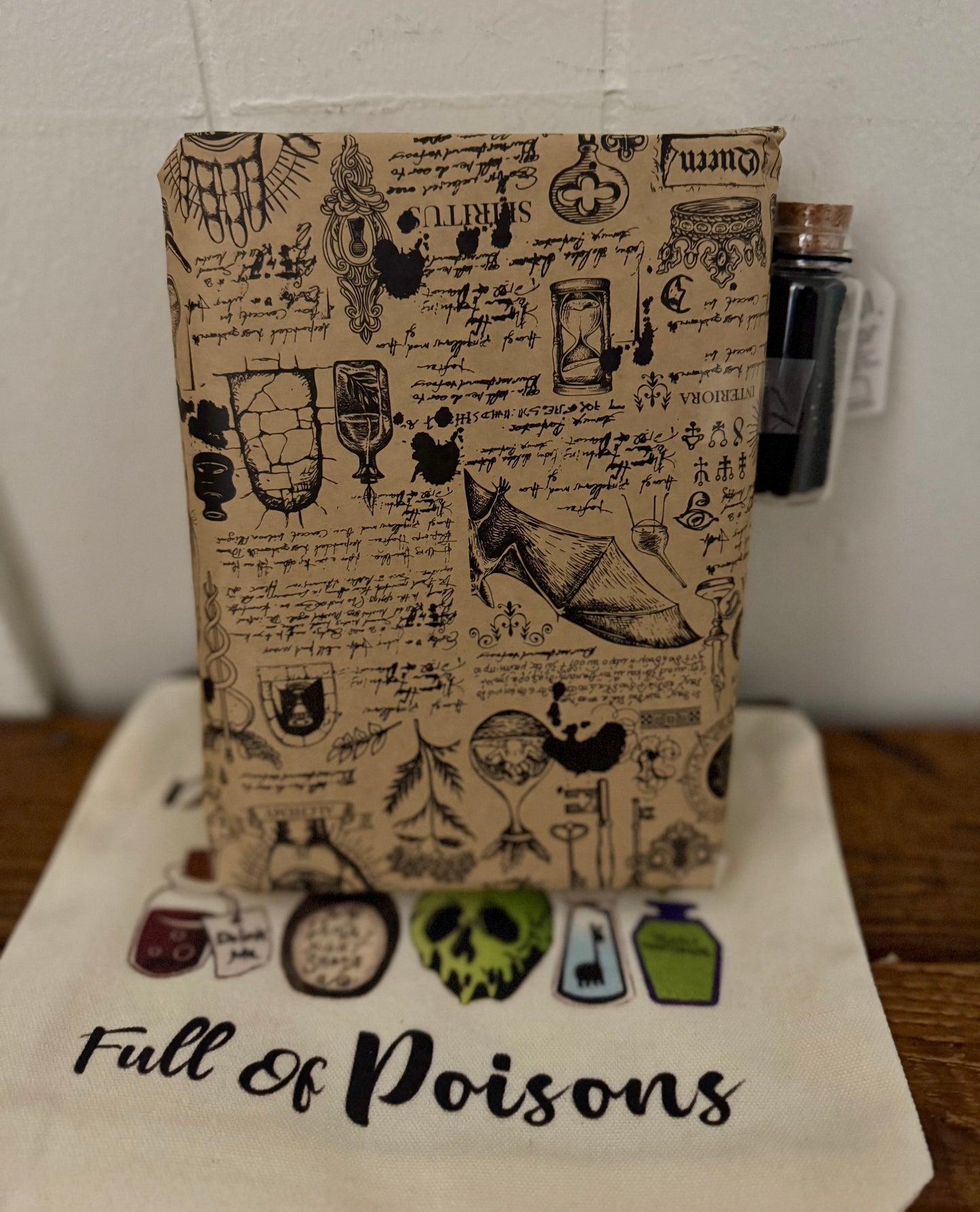 Notebook with vintage-style poison bottle and skull design on a white surface with 'Full of Poisons' text.