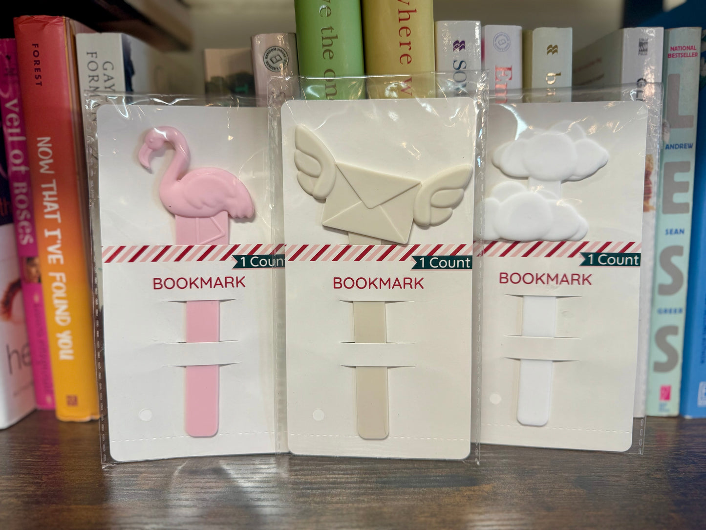 Three bookmarks in packaging with a pink flamingo, beige envelope, and white sheep design on a bookshelf background.