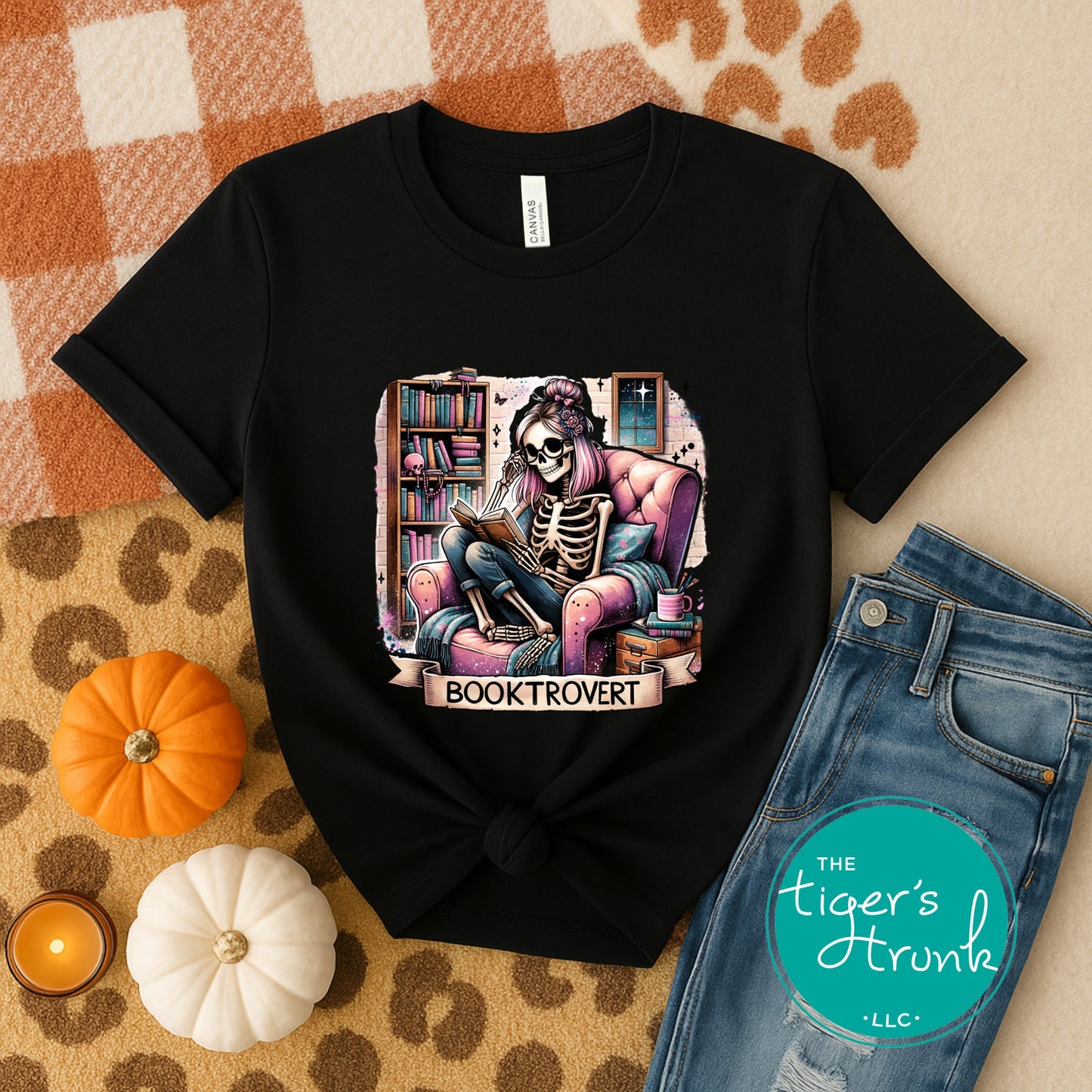 Black t-shirt with skeleton reading a book design, placed on a checkered surface with pumpkins and jeans.