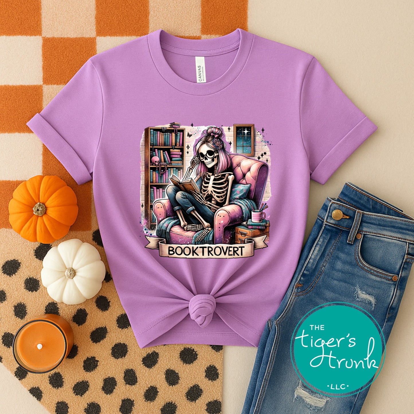 Purple t-shirt with skeleton reading a book design, placed on a checkered fabric background with pumpkins and jeans.