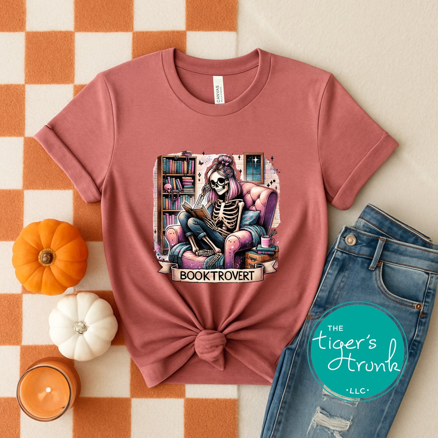 Pink t-shirt with skeleton reading a book design, placed on an orange and white checkered surface with jeans and pumpkins.