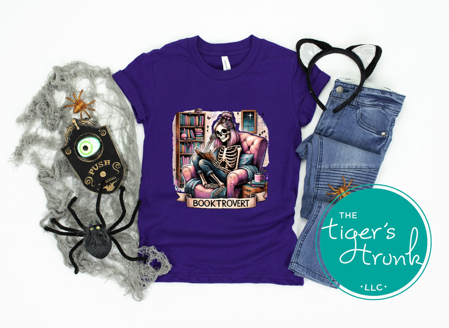 Purple t-shirt with skeleton design, spider decorations, and jeans on a white background with 'The Tiger's Trunk' logo.