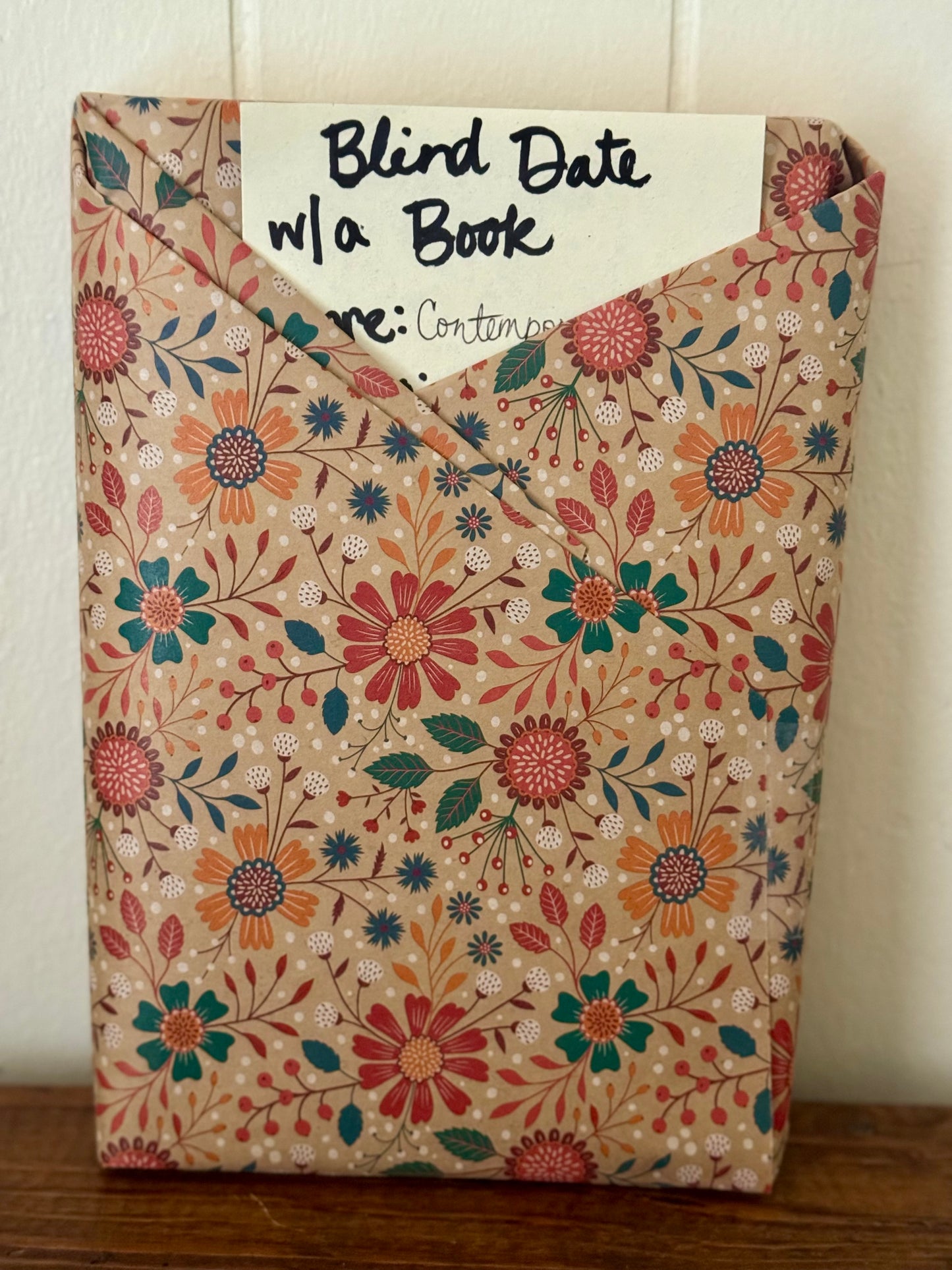 Floral-patterned book with a card labeled 'Blind Date w/ a Book' on a wooden surface.