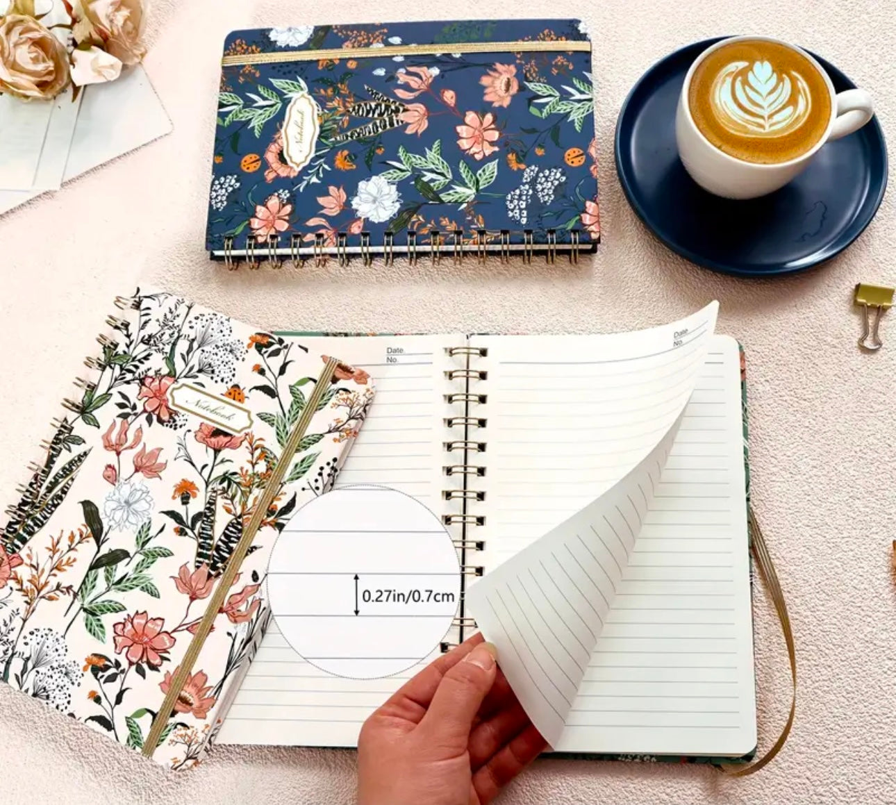 Floral spiral notebook with lined pages and elastic band closure shown open on a neutral surface, styled with a blue coffee cup, matching planner, and gold office accessories in a cozy flat lay setting.
