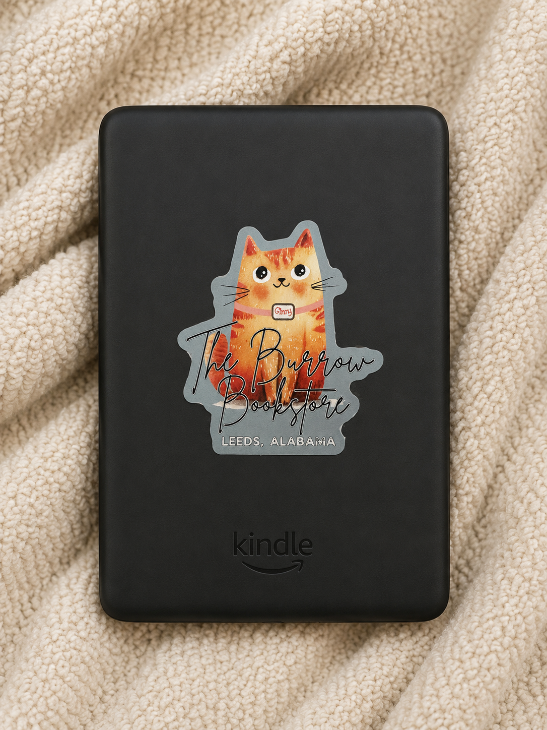 Sage green Ginny the bookstore cat die cut sticker featuring The Burrow Bookstore logo displayed on a Kindle resting on a cozy cream blanket.
