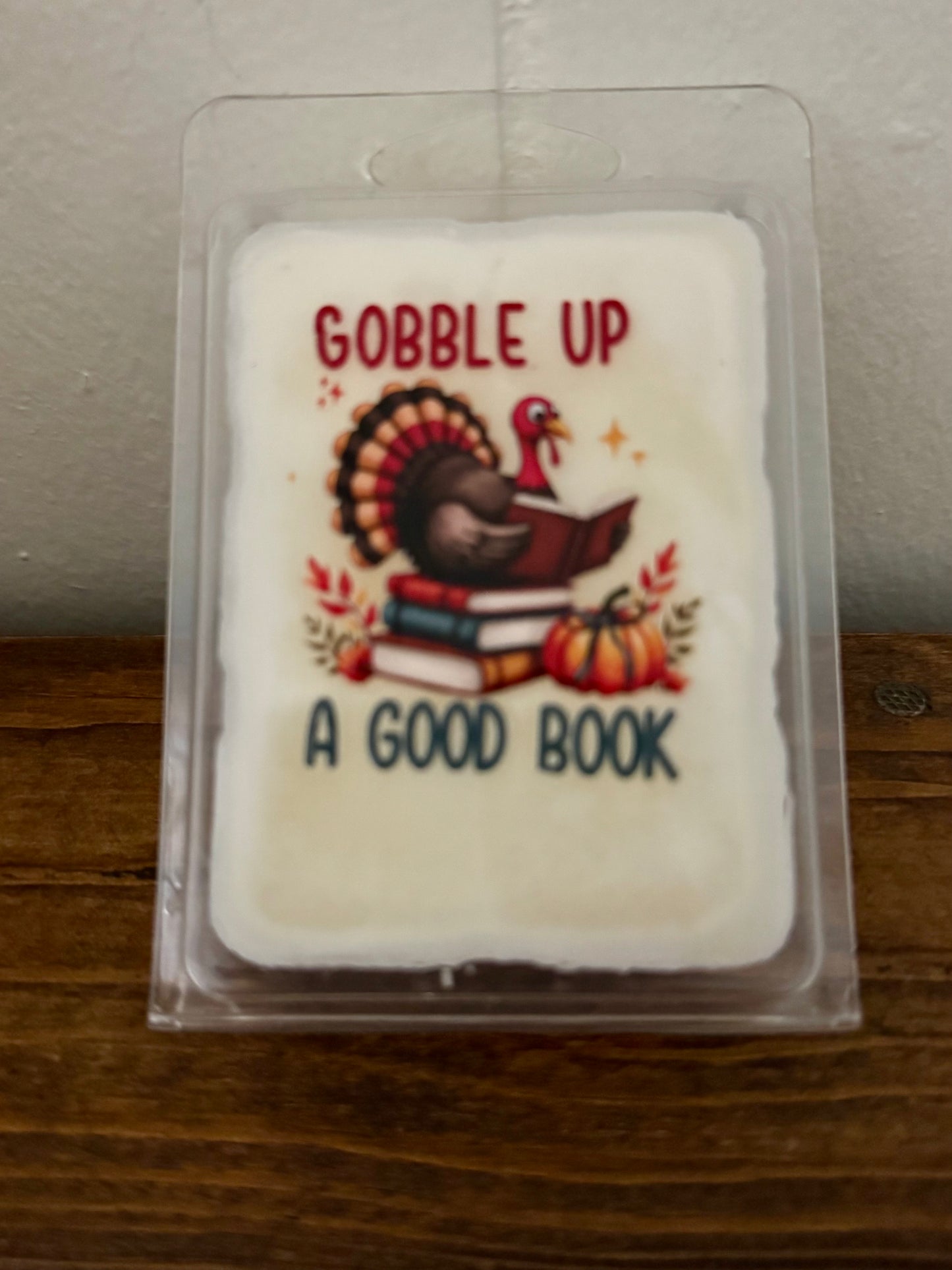 Wax melt with a turkey design and text 'Gobble Up A Good Book' in a clear packaging on a wooden surface.