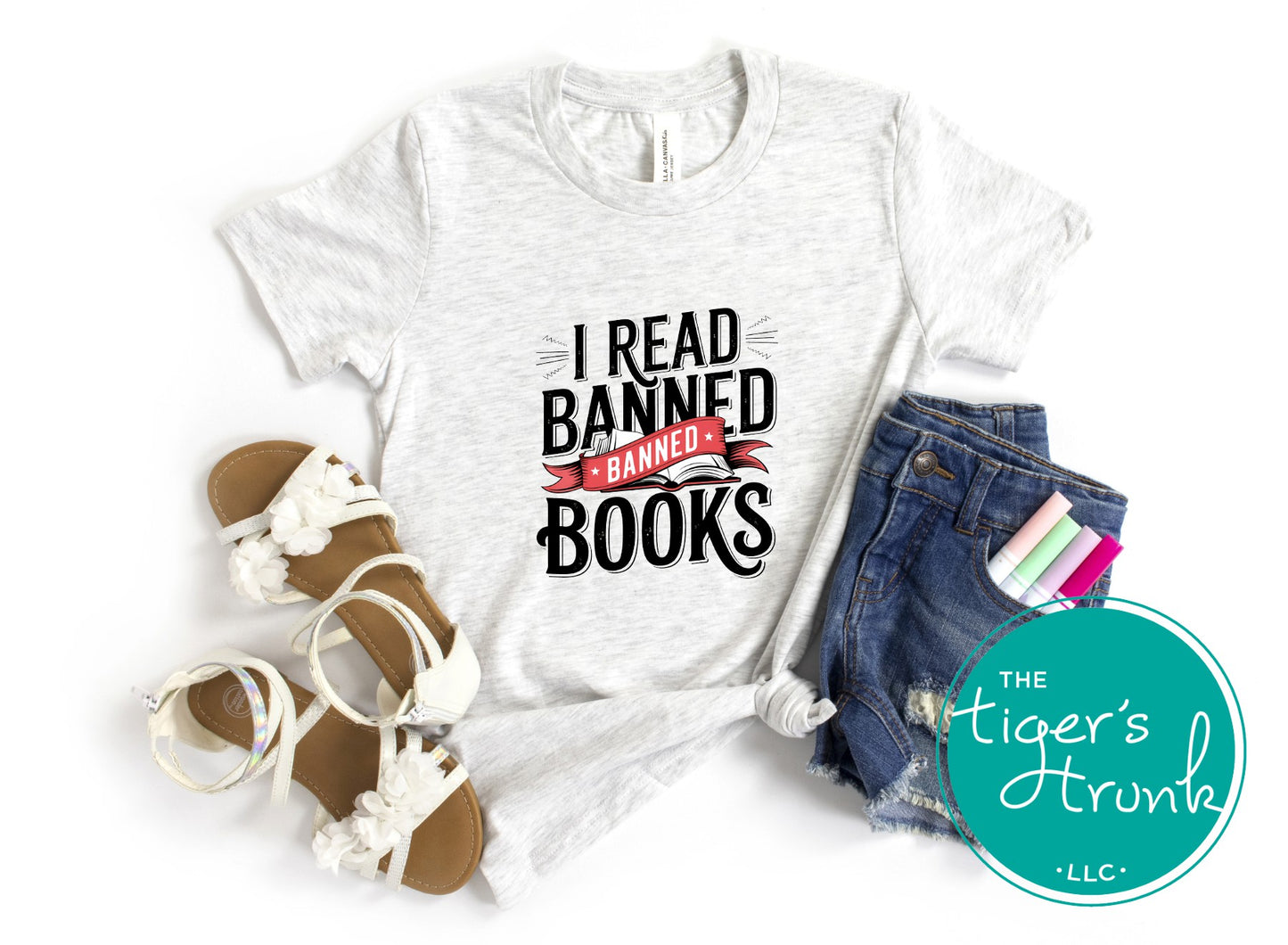 White t-shirt with 'I Read Banned Books' text, sandals, denim shorts, and books on a white background with 'The Tiger's Trunk' logo.