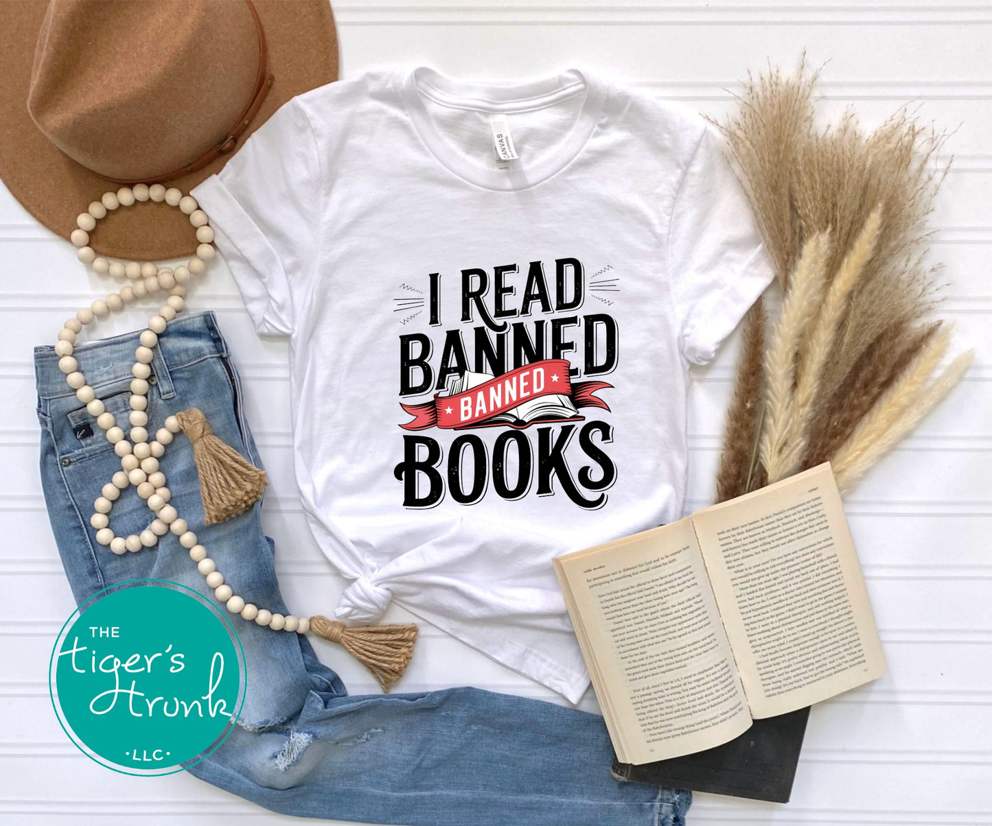 White t-shirt with 'I Read Banned Books' text, surrounded by denim jeans, a hat, and an open book on a white surface.