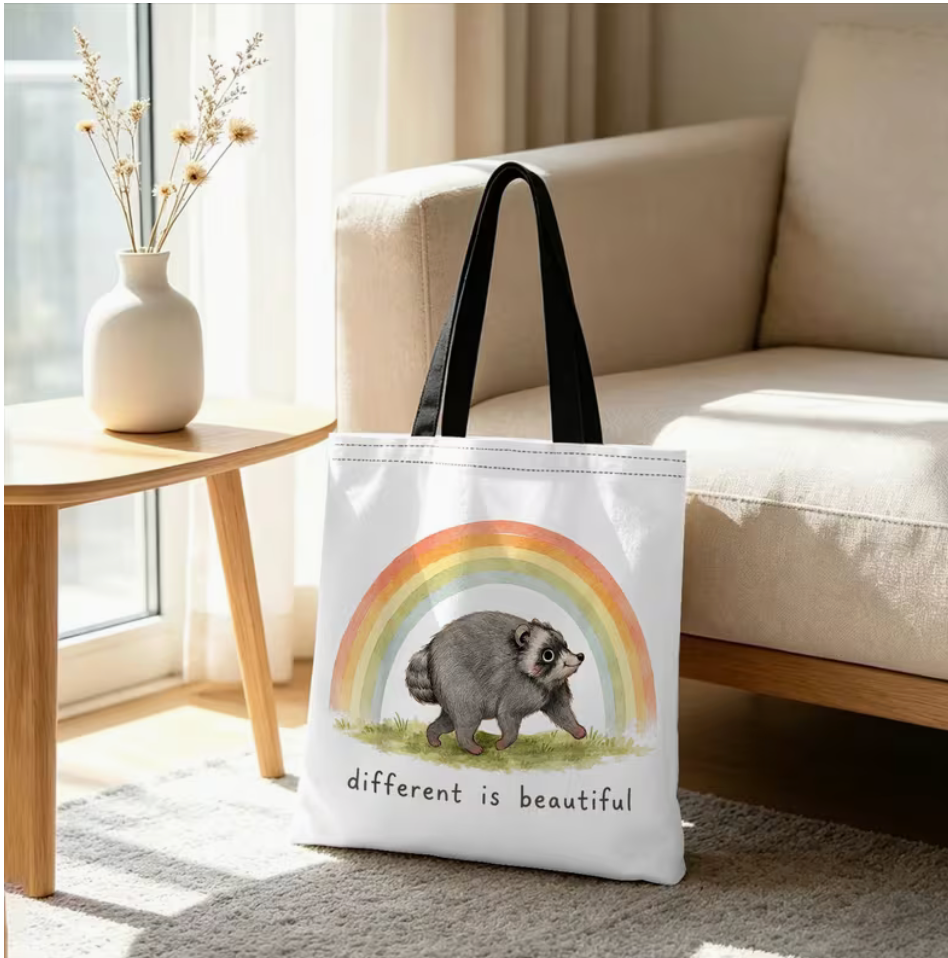 Jimothy the Raccoon Different Is Beautiful tote bag with rainbow design displayed beside a sofa