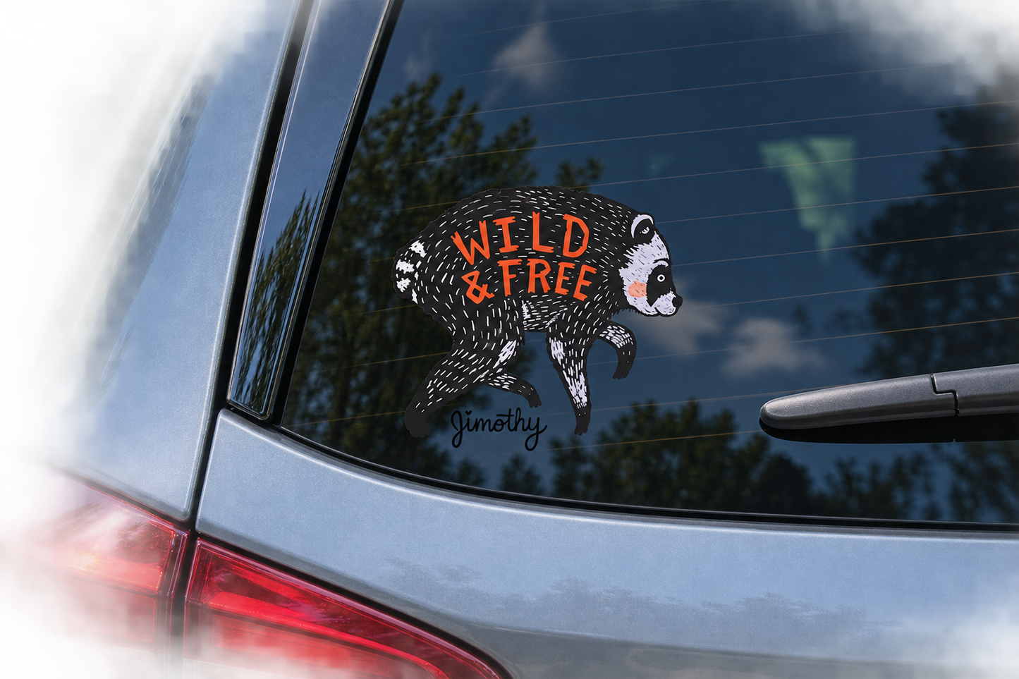 A raccoon-shaped car decal with the words 'WILD & FREE' and a small 'jimothy' signature, applied to the rear window of a silver car.