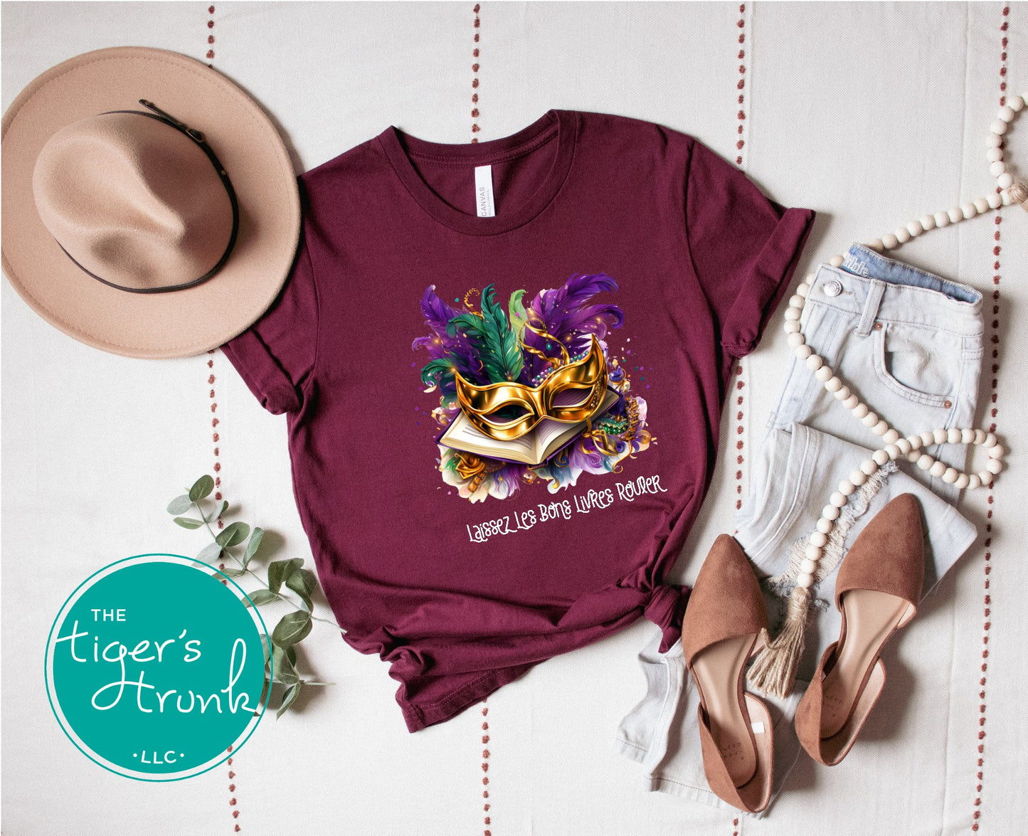 Maroon t-shirt with Mardi Gras-themed design, hat, shoes, and jeans on a white background.