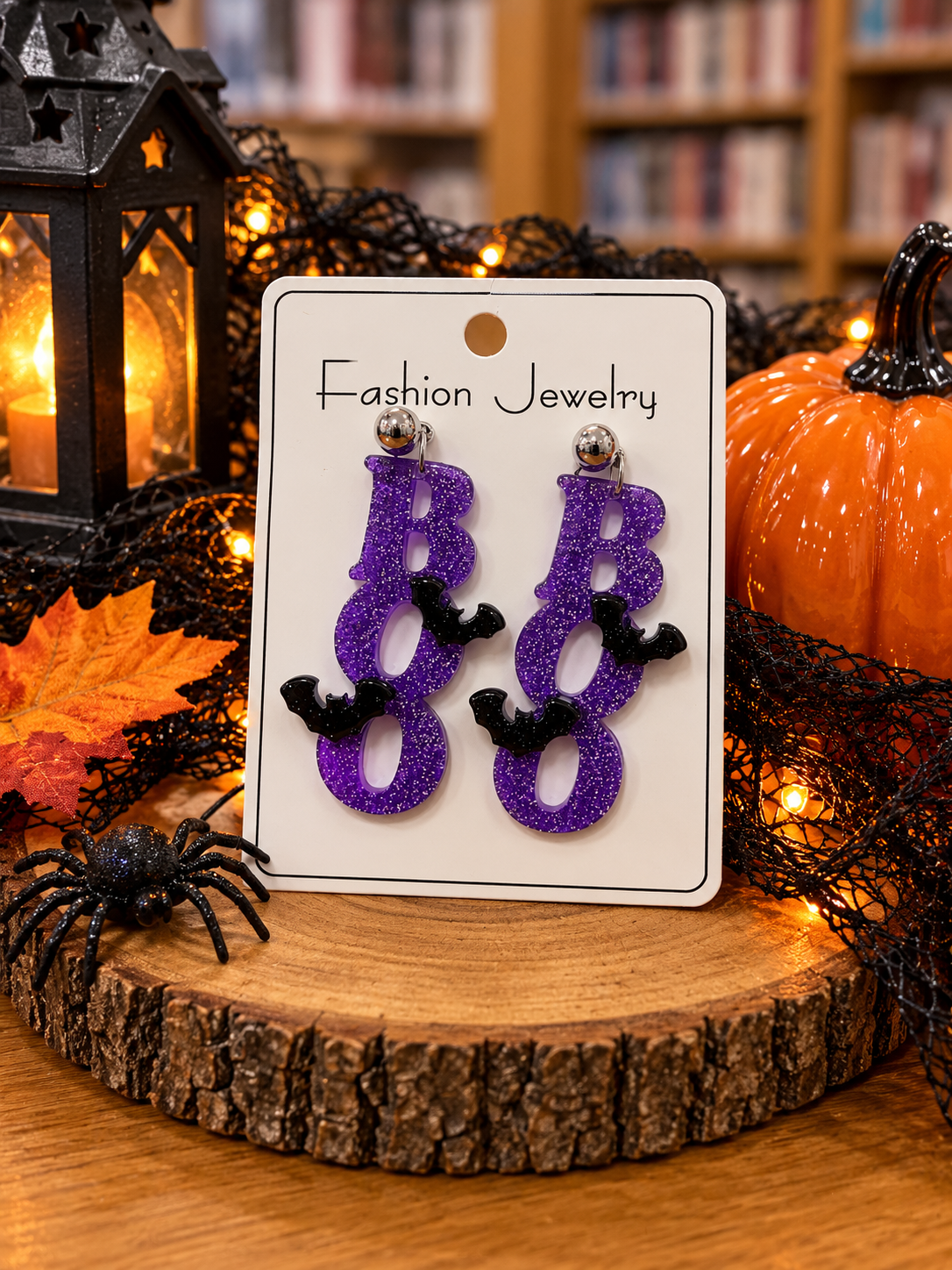 Purple glitter BOO Halloween dangle earrings featuring vertically stacked letters, black bat accents, and silver-tone ball stud posts displayed on a white jewelry card surrounded by festive Halloween decorations.