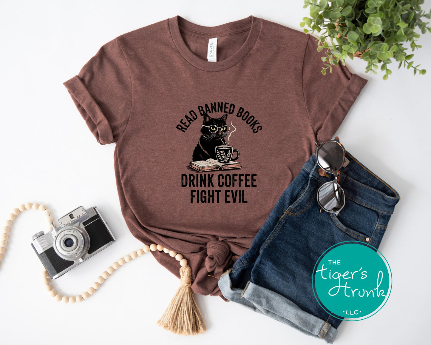 Brown t-shirt with a cat graphic and text, placed on a white surface with jeans, camera, and plant.