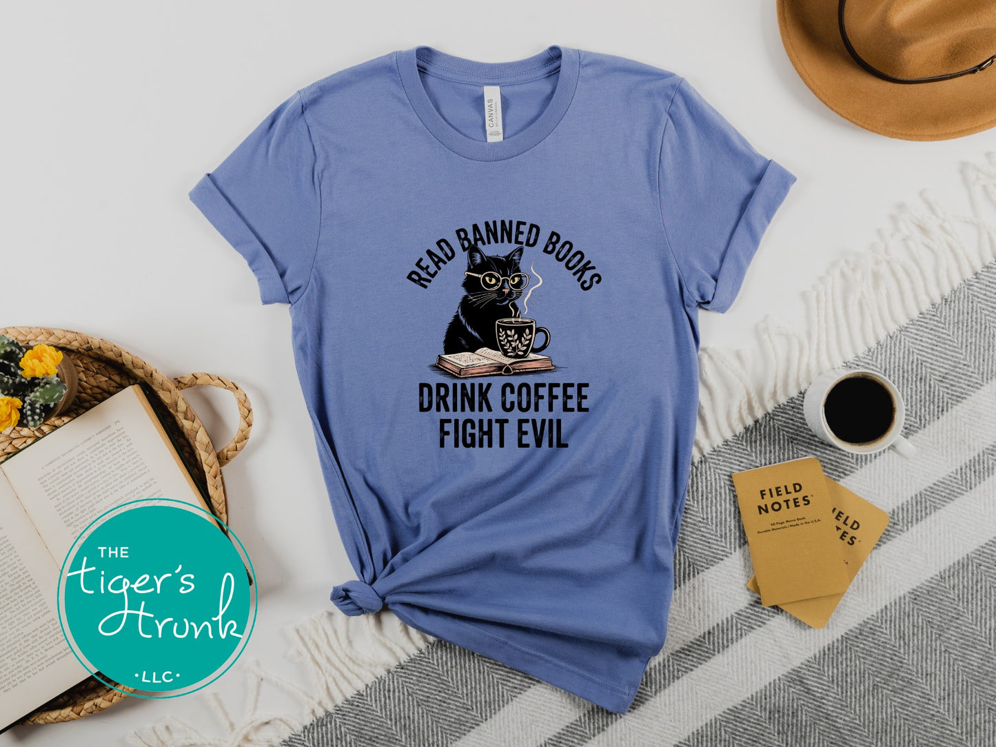 Blue t-shirt with a cat graphic and text on a white surface with a hat, book, and coffee cup.