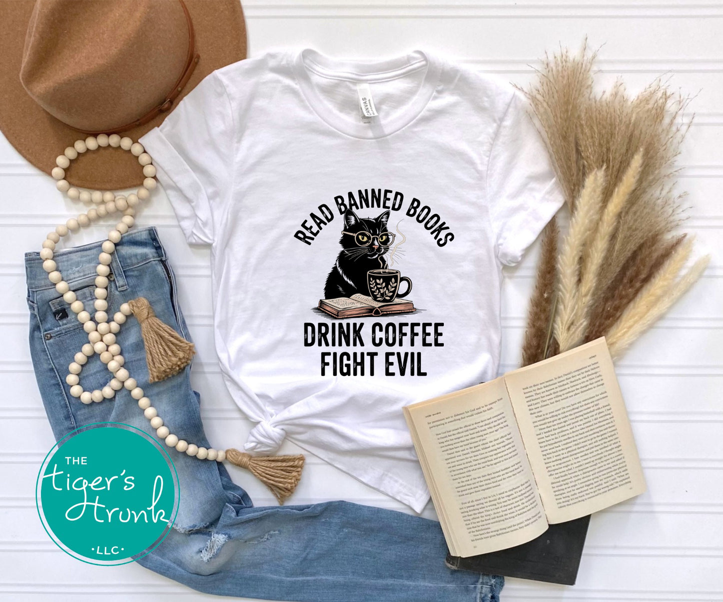 White t-shirt with a black cat graphic and text, placed on a white surface with jeans, a hat, and an open book.