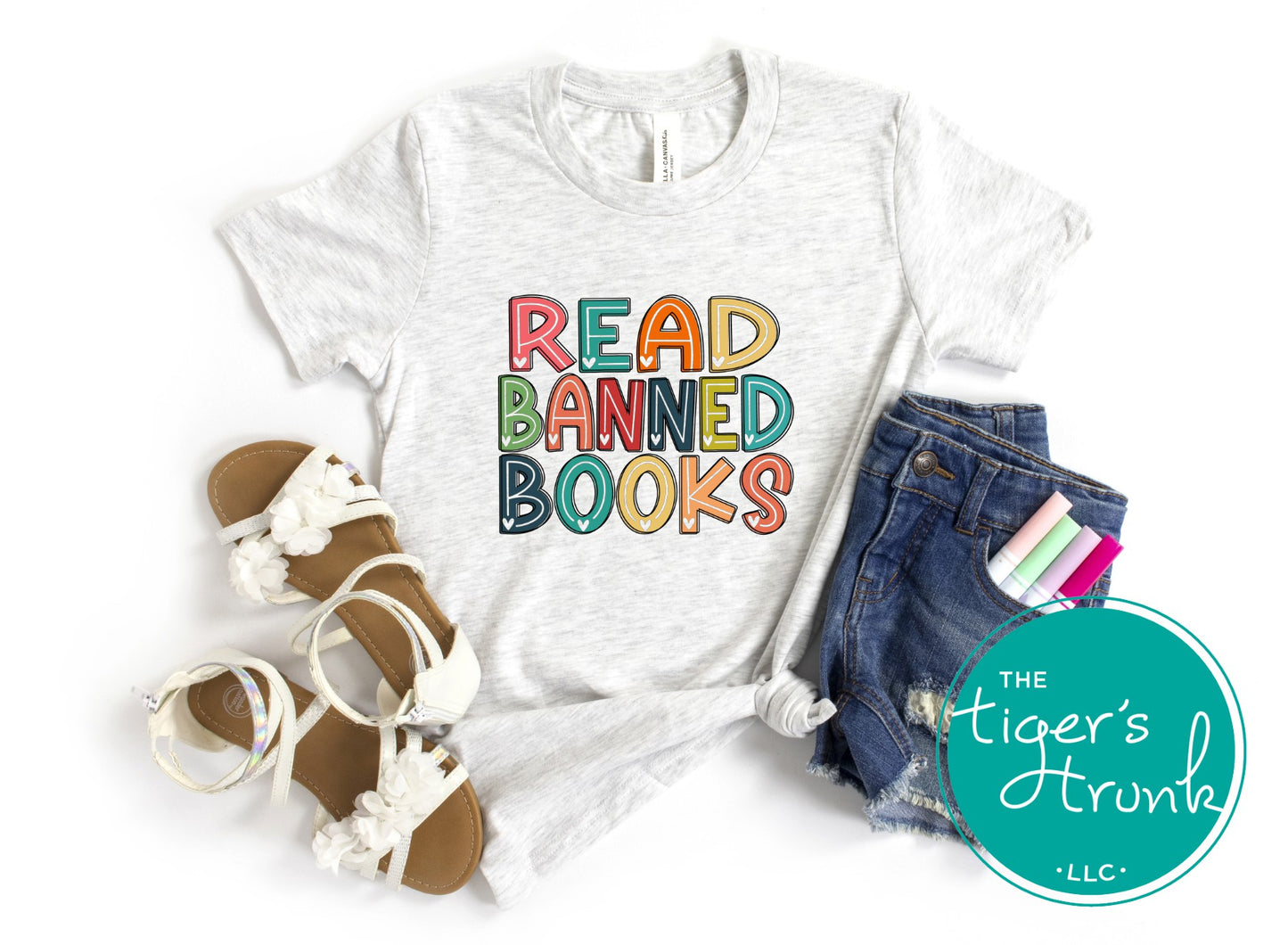 White t-shirt with 'READ BANNED BOOKS' text, sandals, denim shorts, and stationery on a white background. Brand logo 'The Tiger's Trunk' visible.