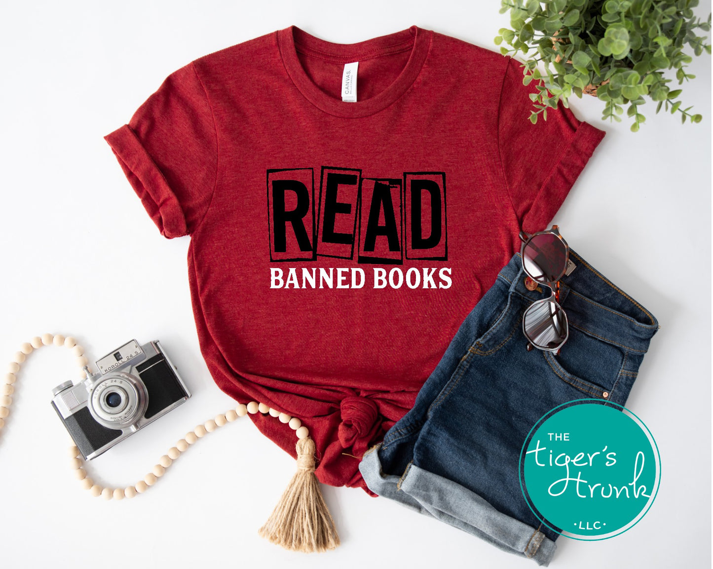 Red t-shirt with 'READ BANNED BOOKS' text, sunglasses, camera, and denim shorts on a white background.
