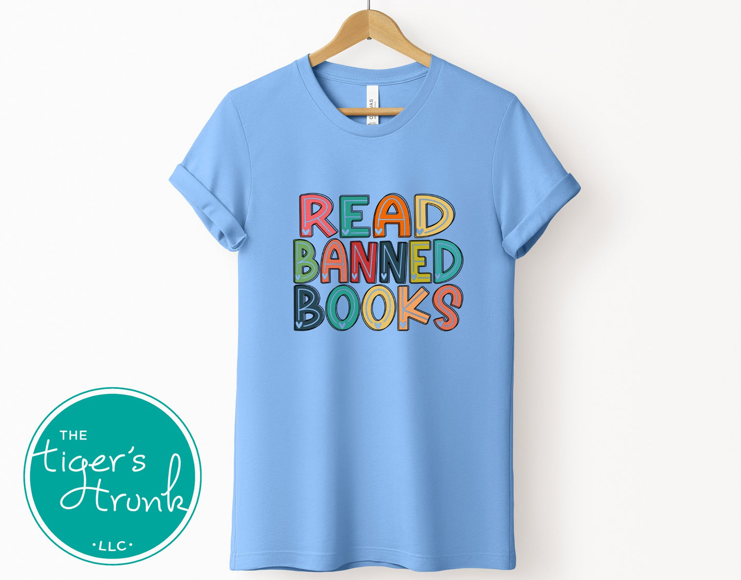 Light blue t-shirt with colorful text 'READ BANNED BOOKS' on a white background, featuring The Tiger's Den logo.