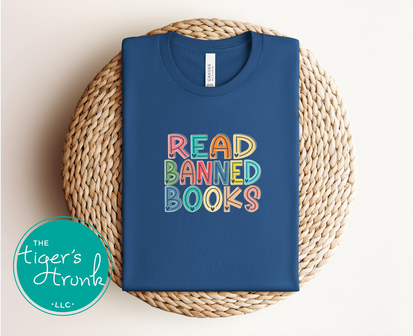 Blue t-shirt with 'READ BANNED BOOKS' text on a woven mat, featuring The Tiger's Trunk LLC logo.
