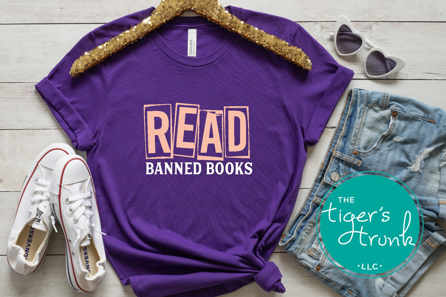 Purple t-shirt with 'READ BANNED BOOKS' text, sunglasses, jeans, and sneakers on a wooden surface with 'The Tiger's Trunk' logo.