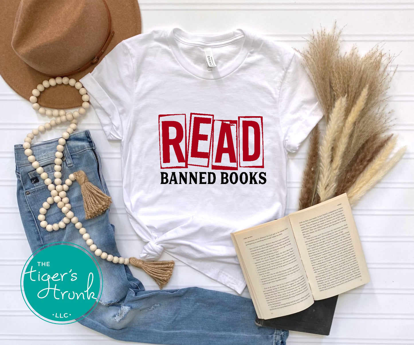 White t-shirt with 'READ BANNED BOOKS' text, surrounded by a hat, book, and decorative items on a white surface.