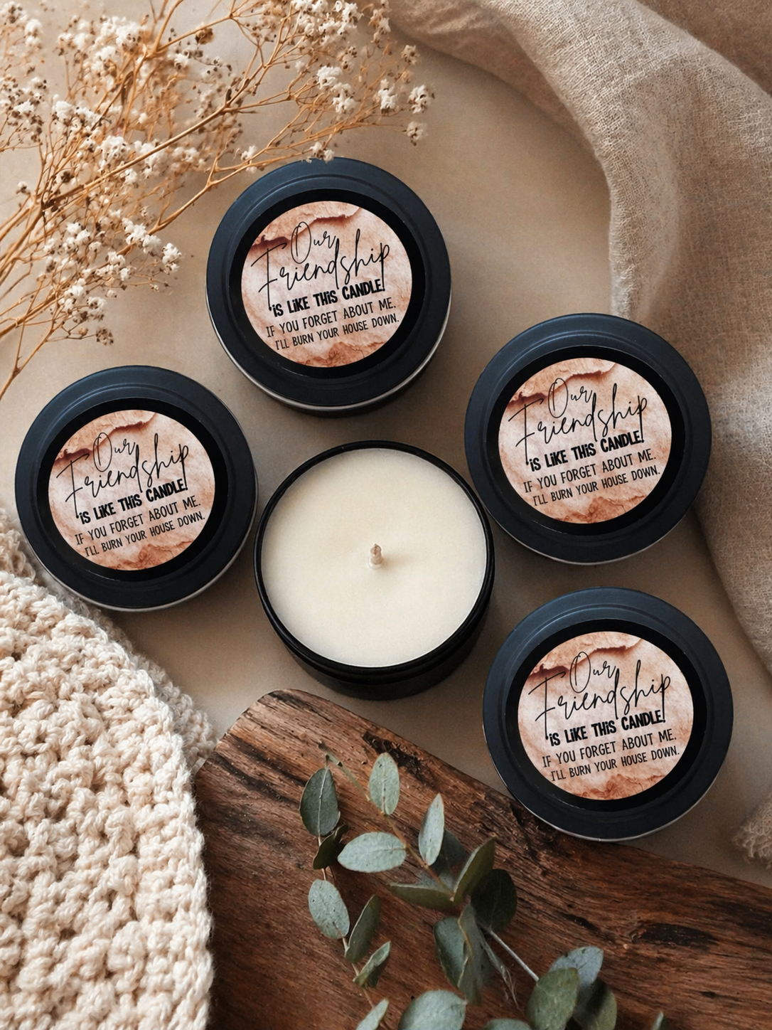 Set of candles with 'My friendship is like this candle' text on a wooden surface with dried flowers and a beige fabric.