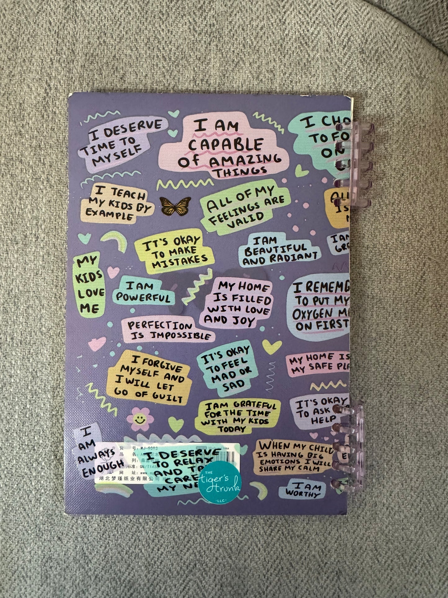 Back cover of a self love journal for moms with colorful affirmation phrases on a purple background, spiral-bound edge visible and Tiger’s Trunk sticker at the bottom.