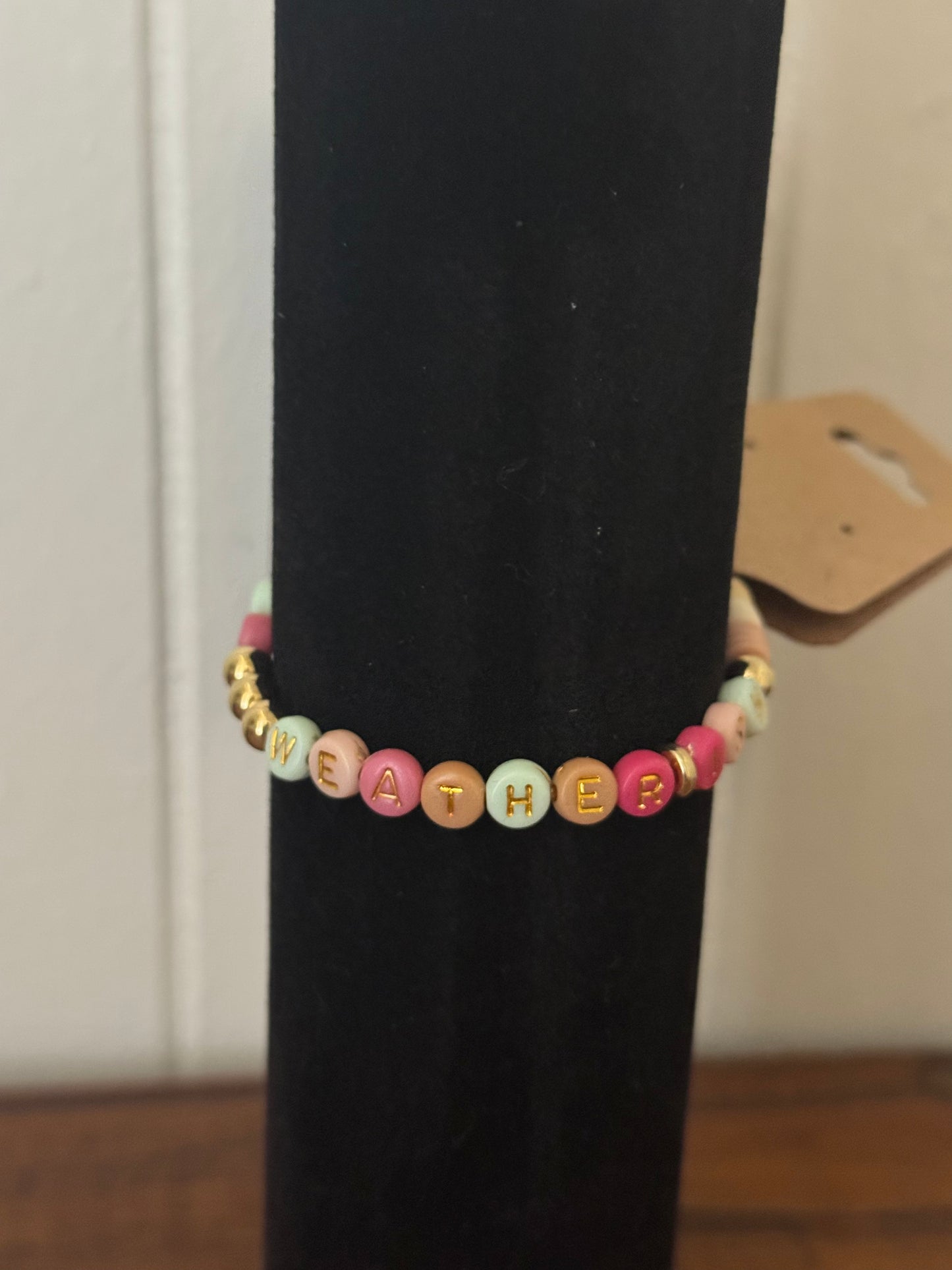 Bracelet with colorful beads spelling 'WEATHER' on a black stand.