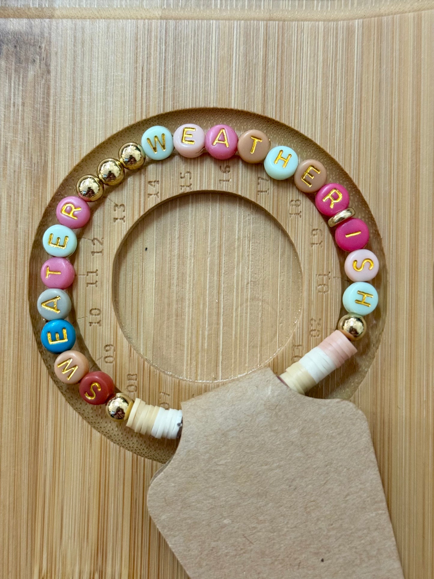 Wooden teething ring with colorful beads spelling 'WEATHER IS' on a wooden background