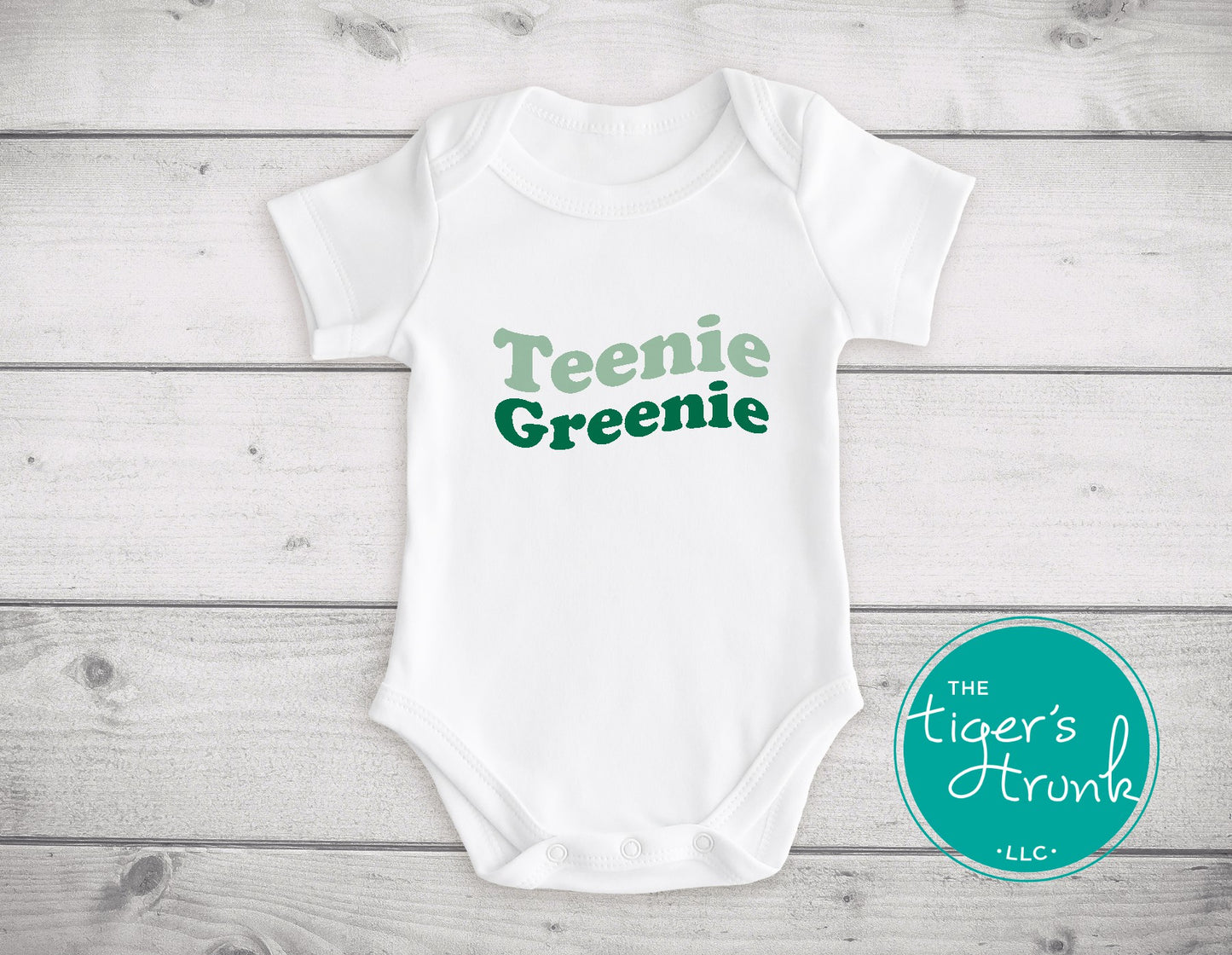 White baby onesie with 'Teenie Greenie' text on a wooden surface, featuring the brand 'The Tiger's Trunk'.