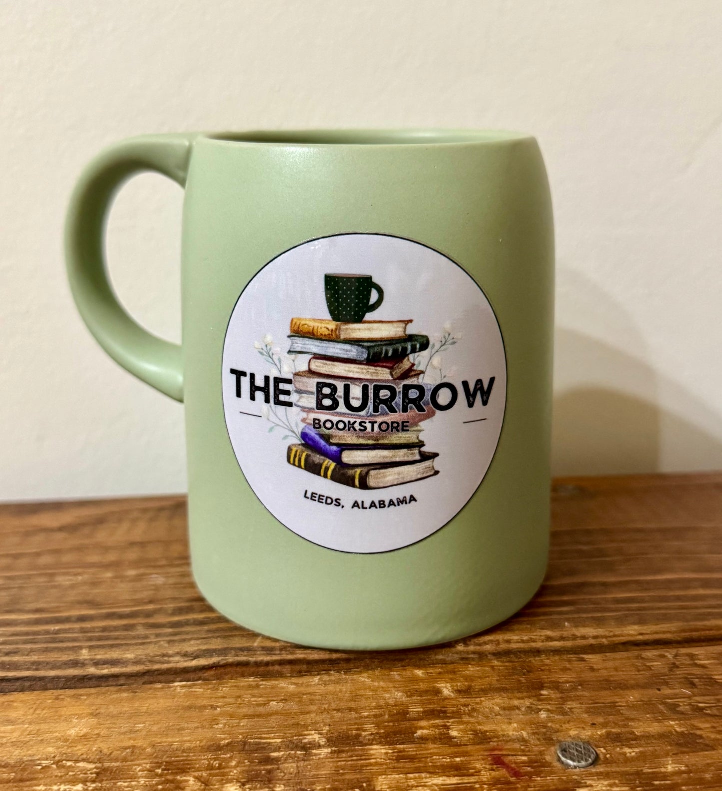 A sage green ceramic mug featuring The Burrow Bookstore logo, perfect for book lovers and independent bookstore supporters. Ideal for coffee, tea, and cozy reading moments.