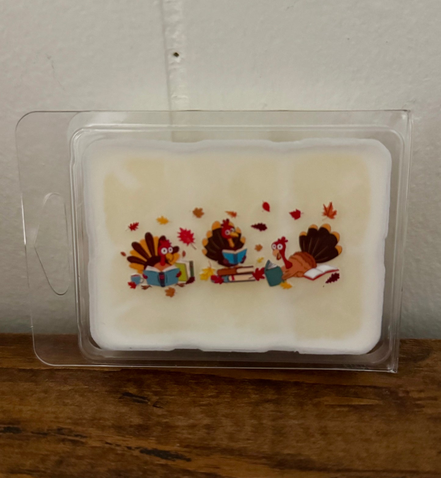 Wax melt with Thanksgiving-themed design of turkeys and leaves in a clear packaging on a wooden surface.