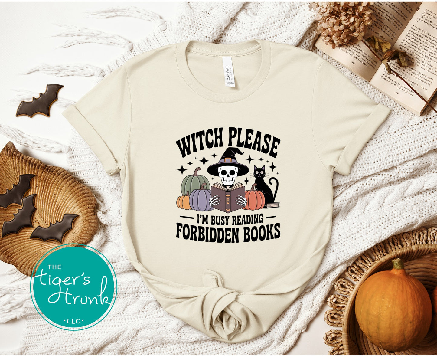 Beige t-shirt with Halloween-themed design and 'Witch Please, I'm Busy Reading Forbidden Books' text, displayed on a textured surface with pumpkins and cookies.