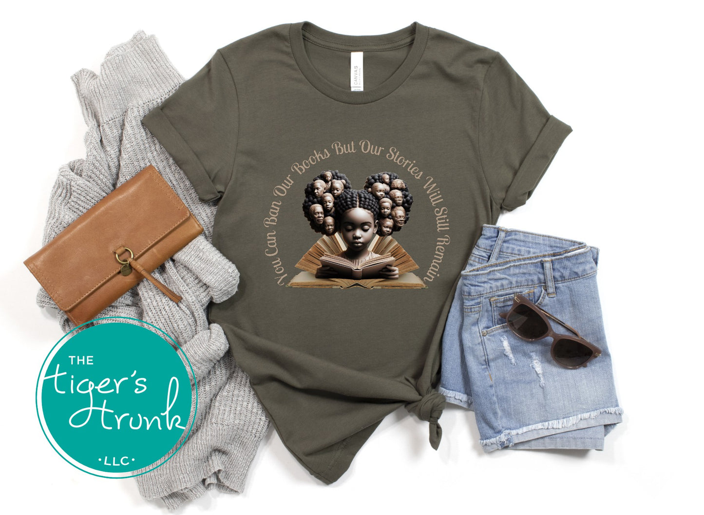 Olive green t-shirt with a graphic design of a woman reading a book, surrounded by a gray sweater, brown wallet, denim shorts, and sunglasses on a white background. The brand 'The Tiger's Trunk' is visible.
