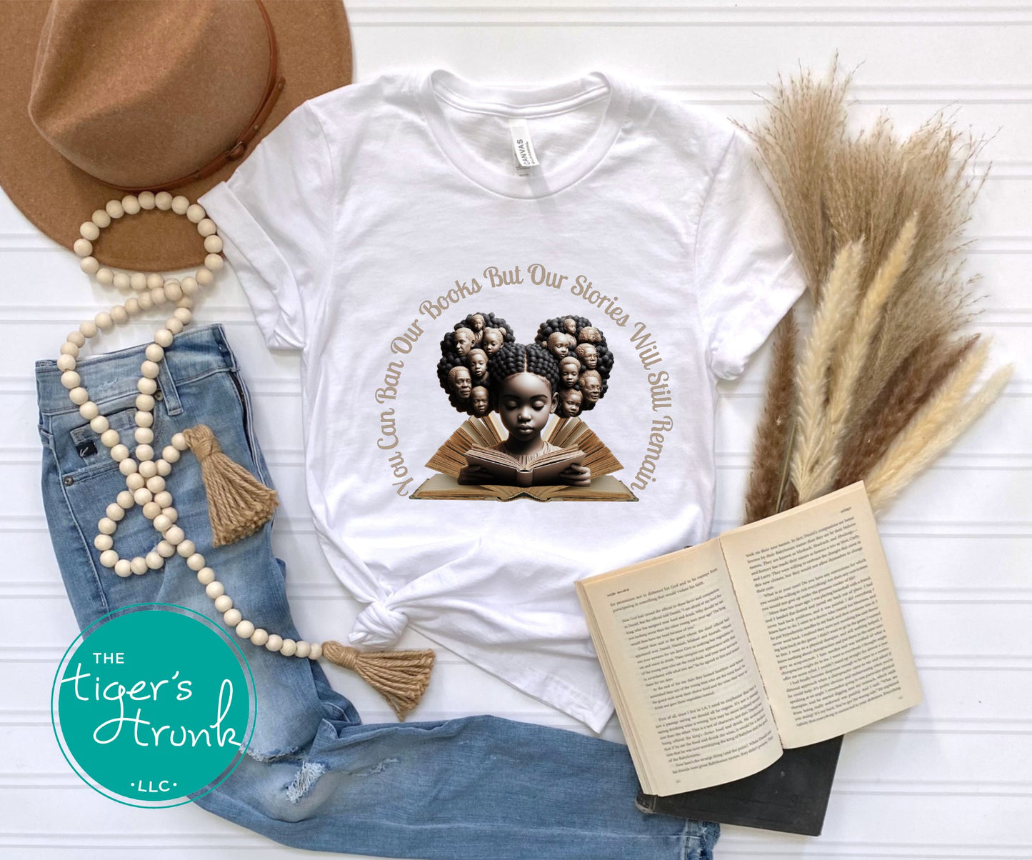 White t-shirt with graphic design featuring a child reading a book, surrounded by denim jeans, an open book, a hat, and decorative elements on a light wooden surface.