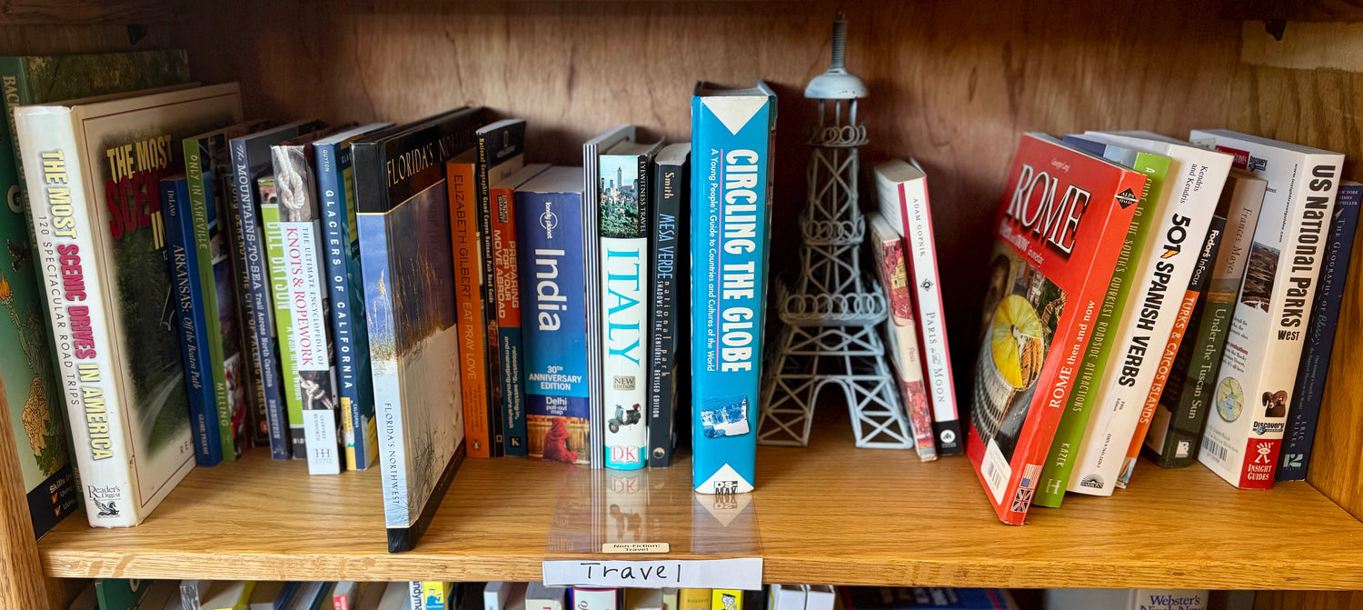 A warm bookstore display featuring travel nonfiction books arranged beside a small globe, postcards, and soft travel-inspired décor.
