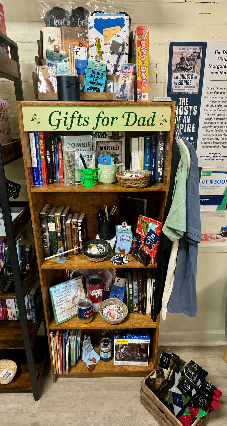 A rustic bookstore table display featuring dad-themed gifts like mugs, candles, shirts, and books, styled with warm, earthy tones and simple, masculine décor.