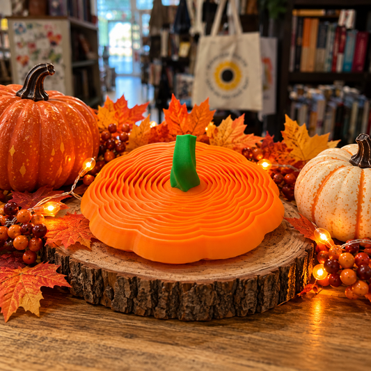 Orange 3D printed pumpkin fidget toy featuring layered concentric rings and a green stem, displayed on a wood slice surrounded by fall leaves, pumpkins, and autumn decorations.