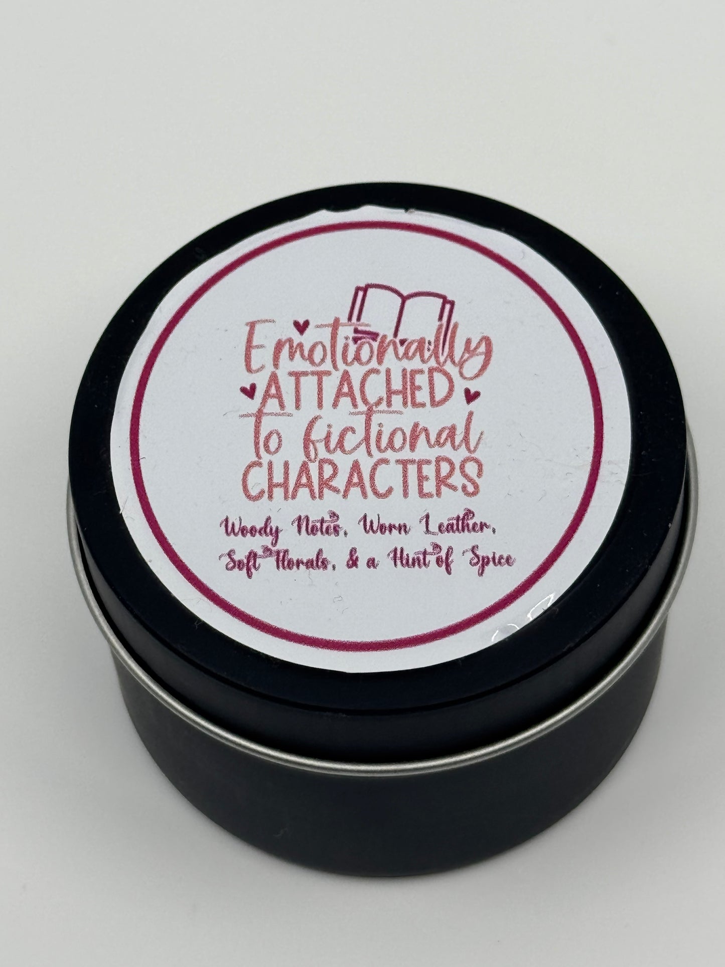 4 oz soy wax candle labeled "Emotionally Attached to Fictional Characters" with a woody, leather, and floral scent, hand-poured with a braided cotton wick.