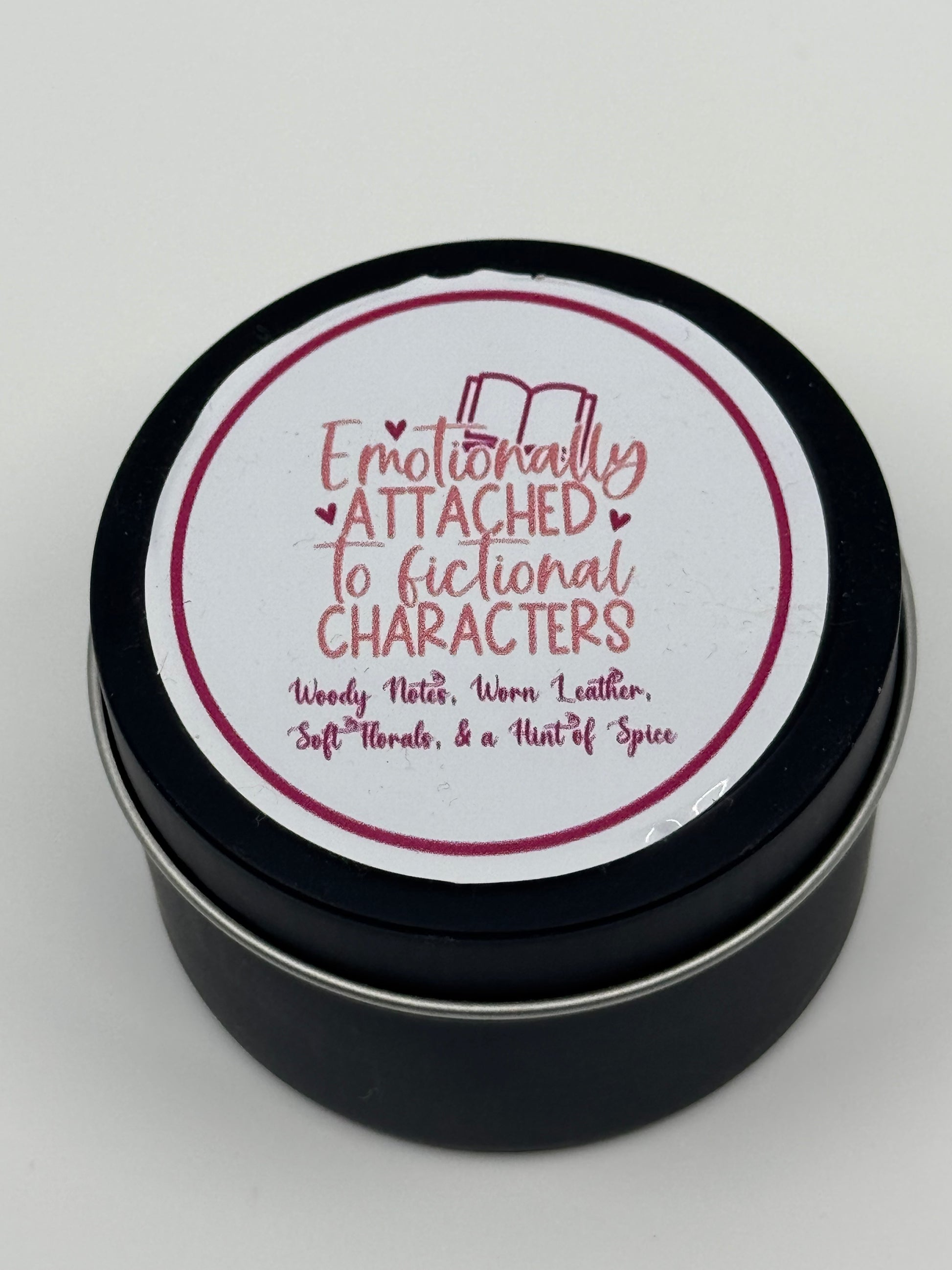 4 oz soy wax candle labeled "Emotionally Attached to Fictional Characters" with a woody, leather, and floral scent, hand-poured with a braided cotton wick.