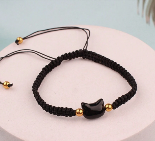 Black macrame bracelet with gold beads and a black cat-shaped charm on a light blue background.