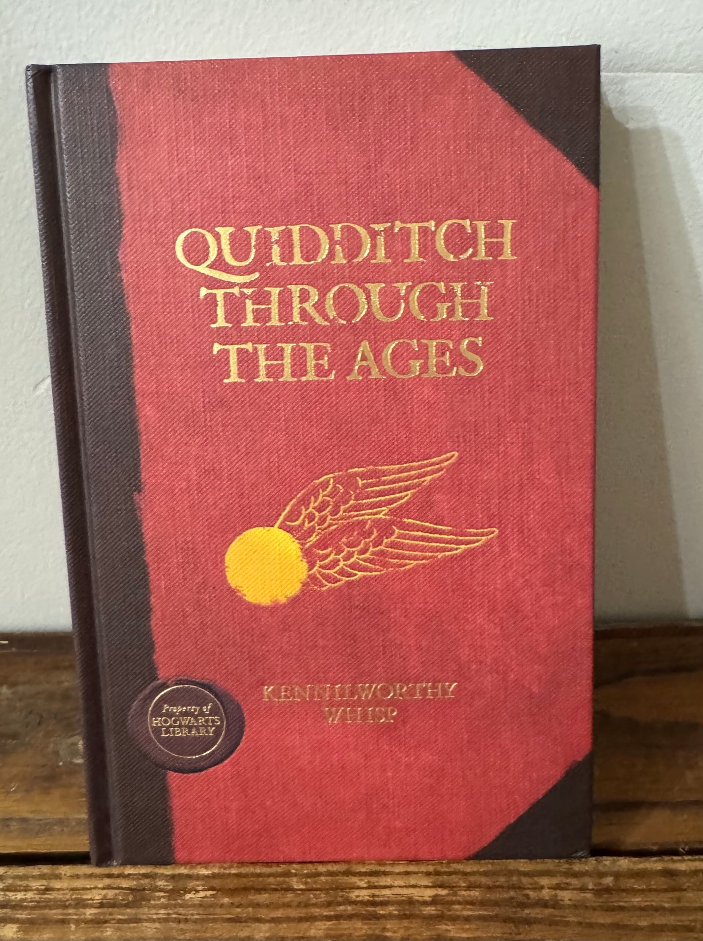 Cover of Quidditch Through the Ages by J.K. Rowling, designed to look like a well-loved library book, featuring a classic emblem of a golden snitch and broomstick on a richly textured background.