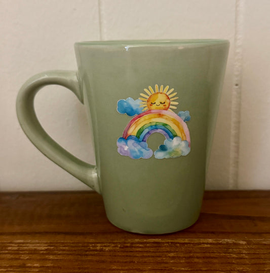Sage green ceramic mug featuring a colorful rainbow and a smiling sunshine design