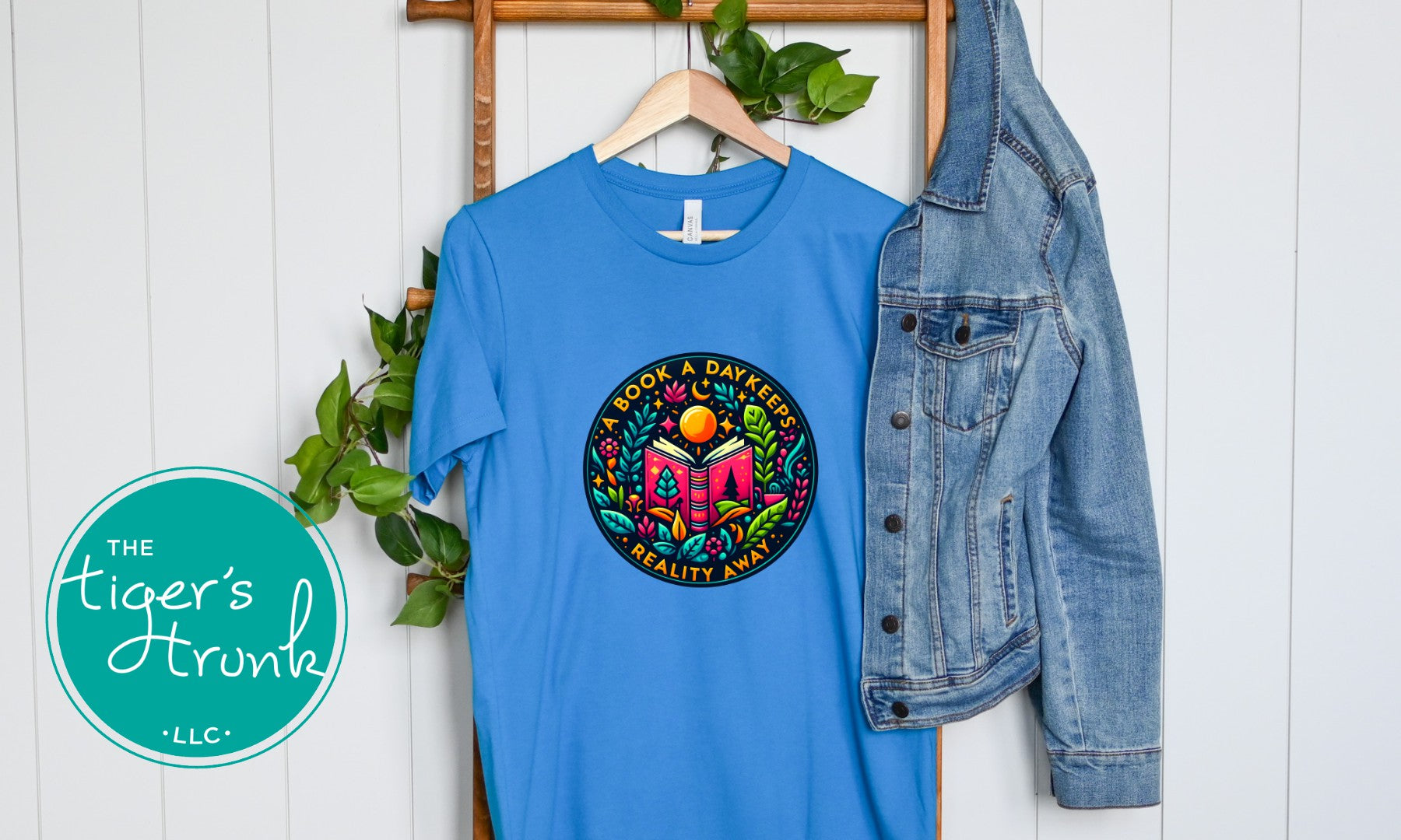 Blue t-shirt with colorful graphic design and denim jacket on a white wooden background, featuring 'The Tiger's Trunk' logo.