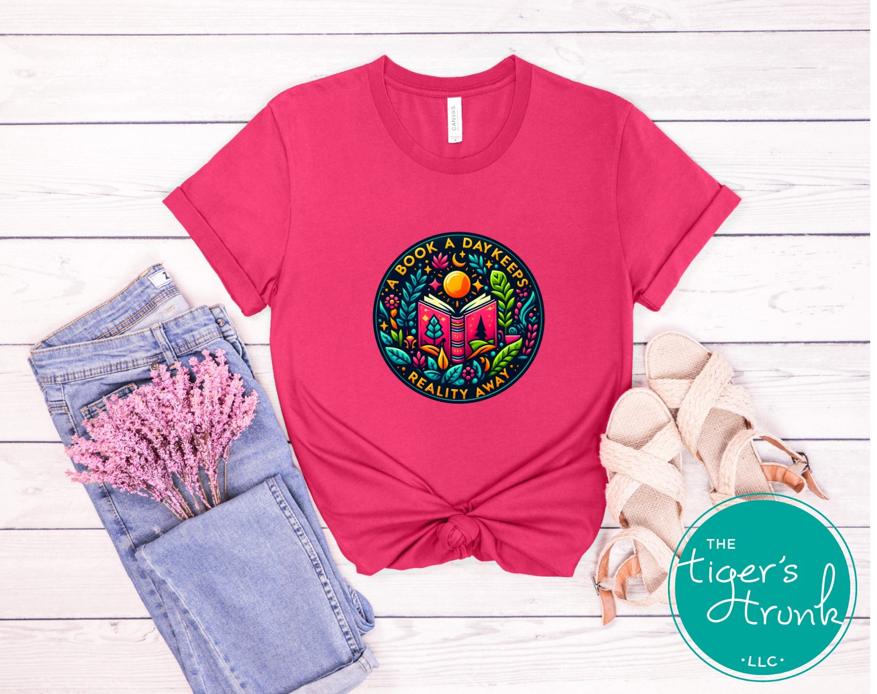 Pink t-shirt with colorful graphic design on a wooden surface, featuring 'The Tiger's Trunk' logo.